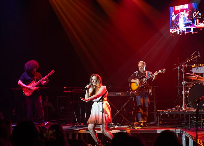 Three members of a band perform on stage, including two men playing guitar and a woman sings into a microphone.